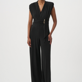 A model wearing a black sleeveless wrap-style jumpsuit with a matching tie belt and a small gold accent, standing against a plain light gray studio background.