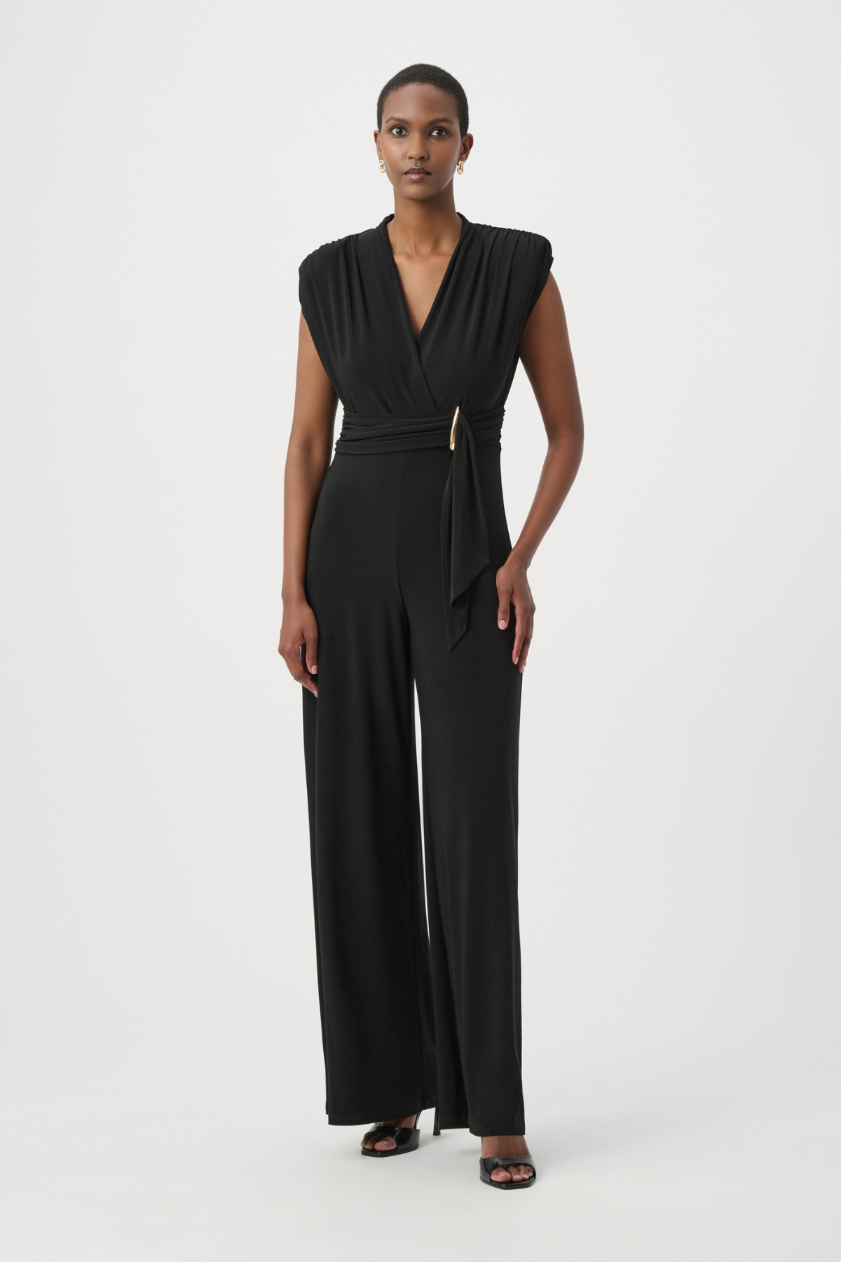 A model wearing a black sleeveless wrap-style jumpsuit with a matching tie belt and a small gold accent, standing against a plain light gray studio background.