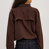 A woman viewed from the back wearing a dark brown long-sleeve cropped top with a wide shoulder yoke and dark blue jeans, against a plain light gray studio background.