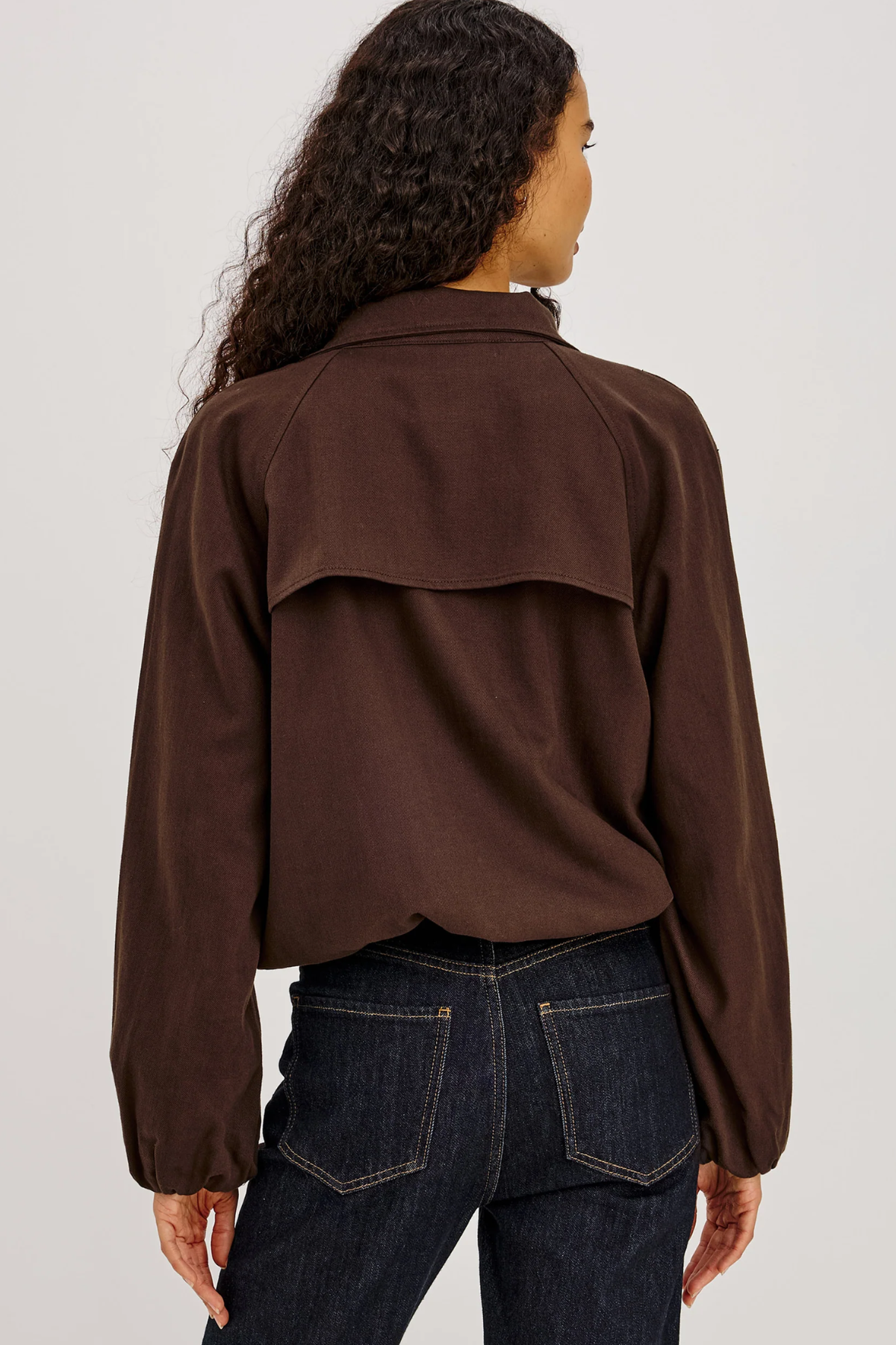 A woman viewed from the back wearing a dark brown long-sleeve cropped top with a wide shoulder yoke and dark blue jeans, against a plain light gray studio background.