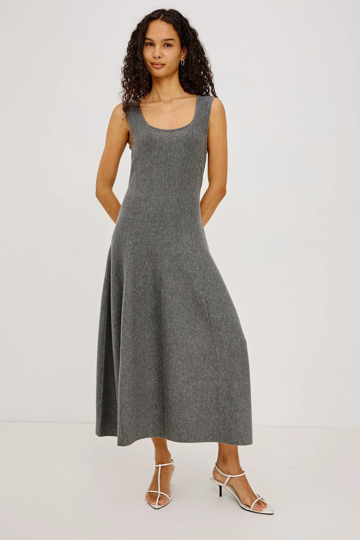 A woman modeling a sleeveless, scoop-neck, midi-length gray knit dress with a flared skirt, paired with white strappy high-heeled sandals, against a plain white studio background.