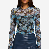 A woman modeling a sheer long-sleeve top with a blue, white, and brown floral pattern, paired with a dark navy ruched skirt, against a light gray background.