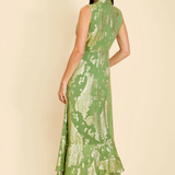A woman modeling a sleeveless green maxi dress with a gold floral pattern, shown from the back against a solid beige background.