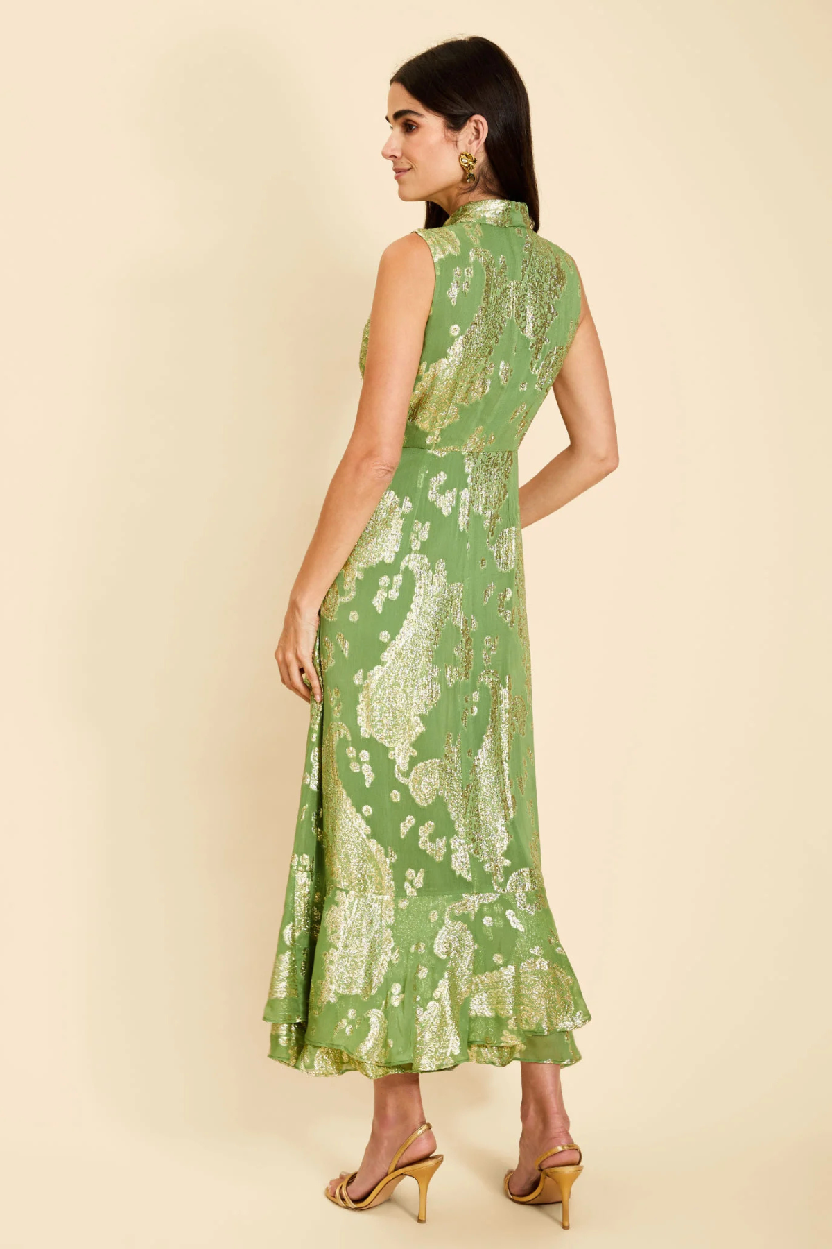 A woman modeling a sleeveless green maxi dress with a gold floral pattern, shown from the back against a solid beige background.