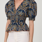 Woman wearing a patterned blue and yellow top with white pants on a plain background