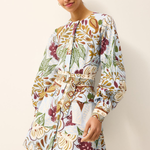 A woman modeling a long-sleeve mini dress with a white base and a multicolor botanical print featuring green, burgundy, and gold tones, set against a plain off-white background.