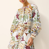 A woman modeling a long-sleeve mini dress with a white base and a multicolor botanical print featuring green, burgundy, and gold tones, set against a plain off-white background.