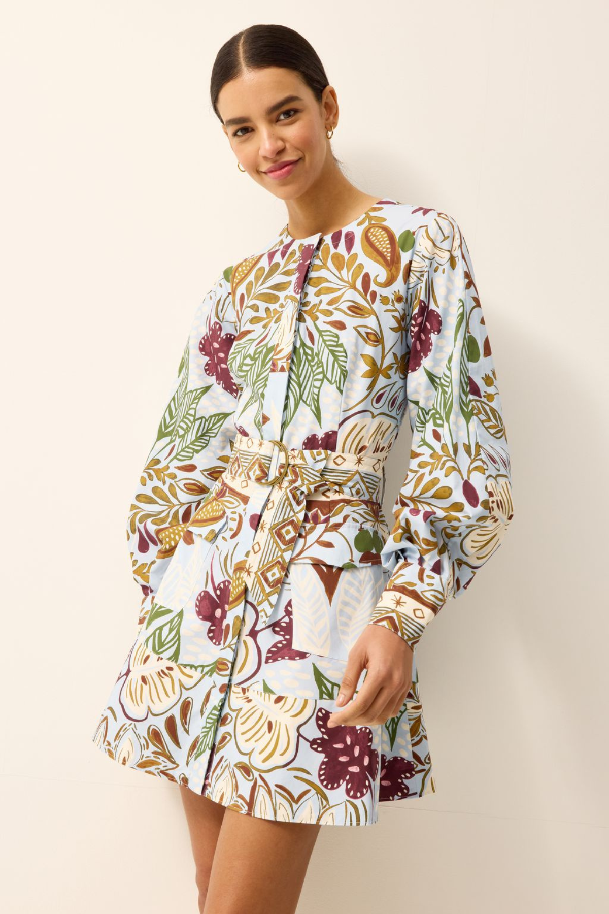 A woman modeling a long-sleeve mini dress with a white base and a multicolor botanical print featuring green, burgundy, and gold tones, set against a plain off-white background.