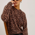 A woman wearing a fuzzy leopard-print knit sweater, dark sunglasses, and black jeans, seated on a wooden chair against a plain light background.