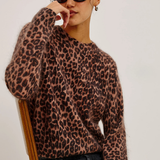 A woman wearing a fuzzy leopard-print knit sweater, dark sunglasses, and black jeans, seated on a wooden chair against a plain light background.