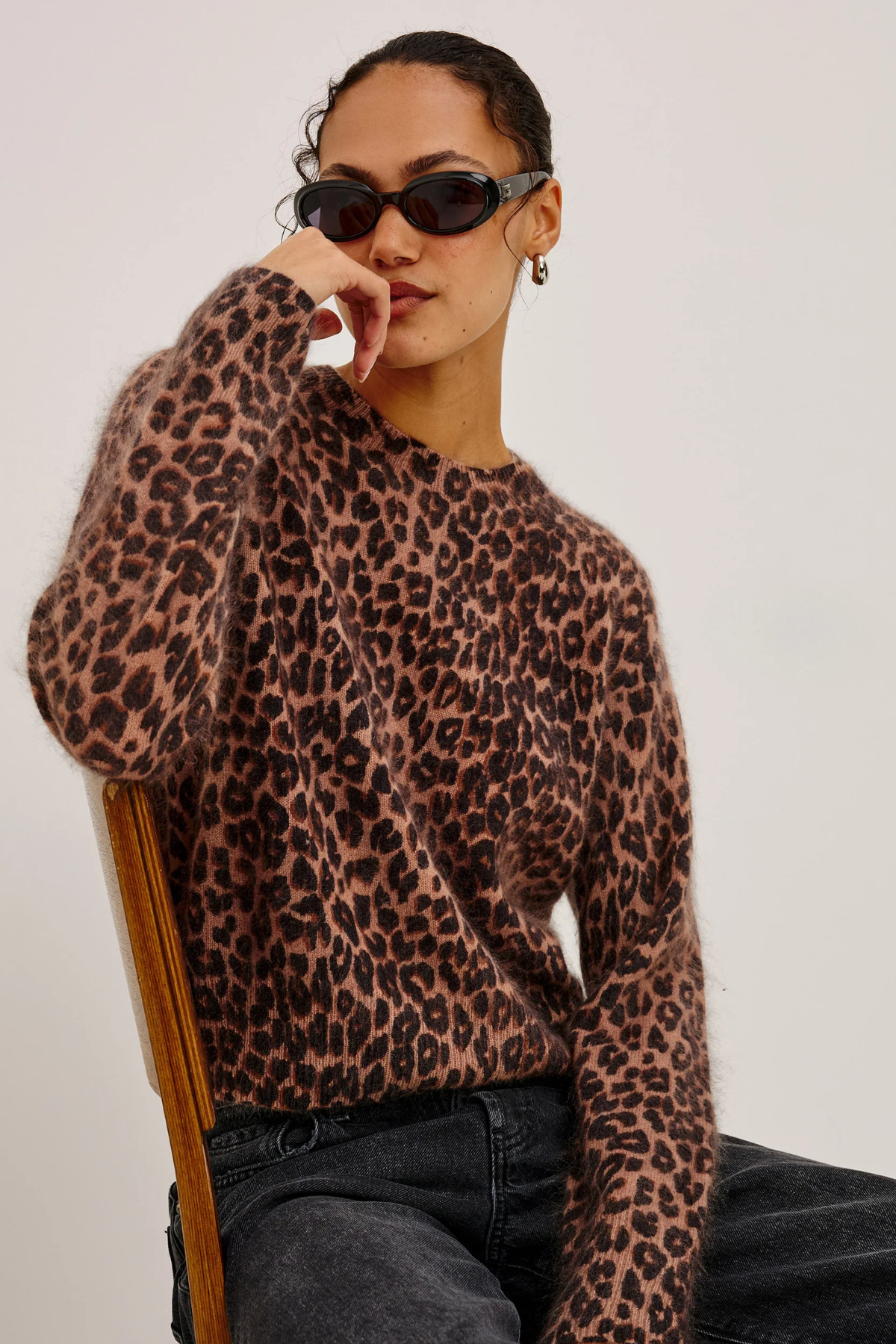 A woman wearing a fuzzy leopard-print knit sweater, dark sunglasses, and black jeans, seated on a wooden chair against a plain light background.