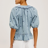 A woman shown from the back wearing a light blue denim top with puff sleeves, a ruffled collar, and small embroidered dots, paired with white pants against a plain light background.