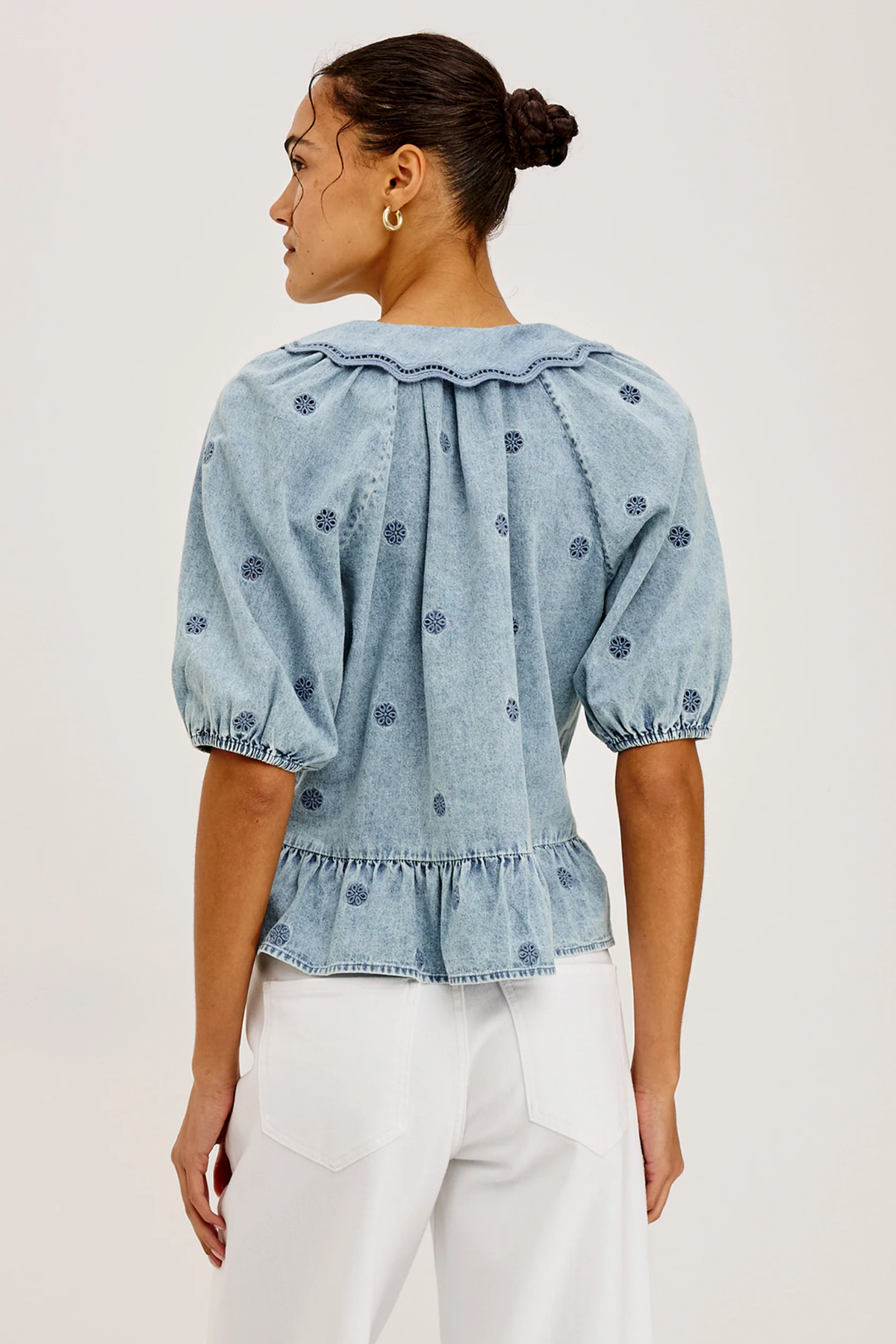 A woman shown from the back wearing a light blue denim top with puff sleeves, a ruffled collar, and small embroidered dots, paired with white pants against a plain light background.