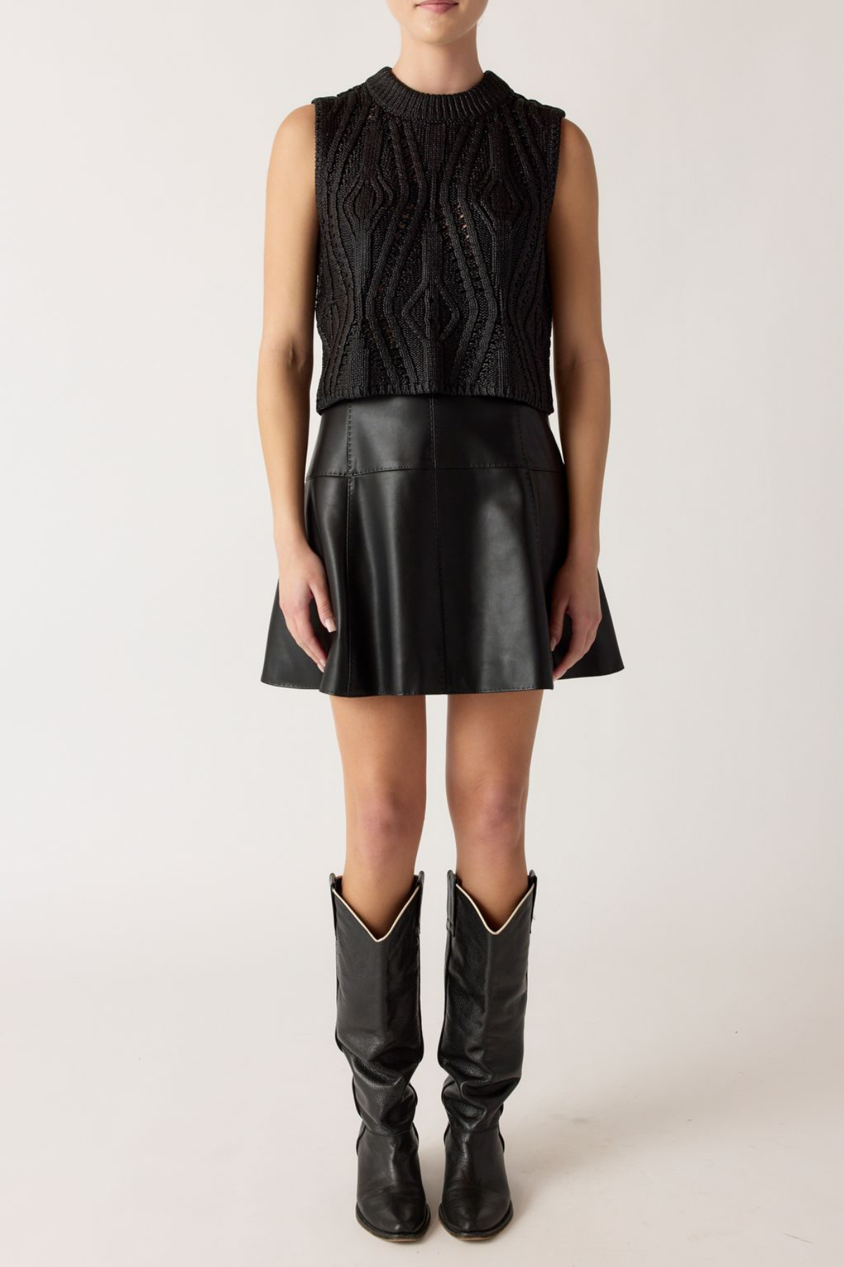 Person wearing a black sleeveless knitted top, black leather skirt, and black cowboy boots on a white background