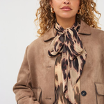 A woman wearing a tan suede jacket, a beige blouse, and a matching leopard-print silk scarf tied around her neck, photographed against a light gray studio background.