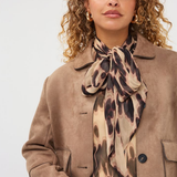 A woman wearing a tan suede jacket, a beige blouse, and a matching leopard-print silk scarf tied around her neck, photographed against a light gray studio background.