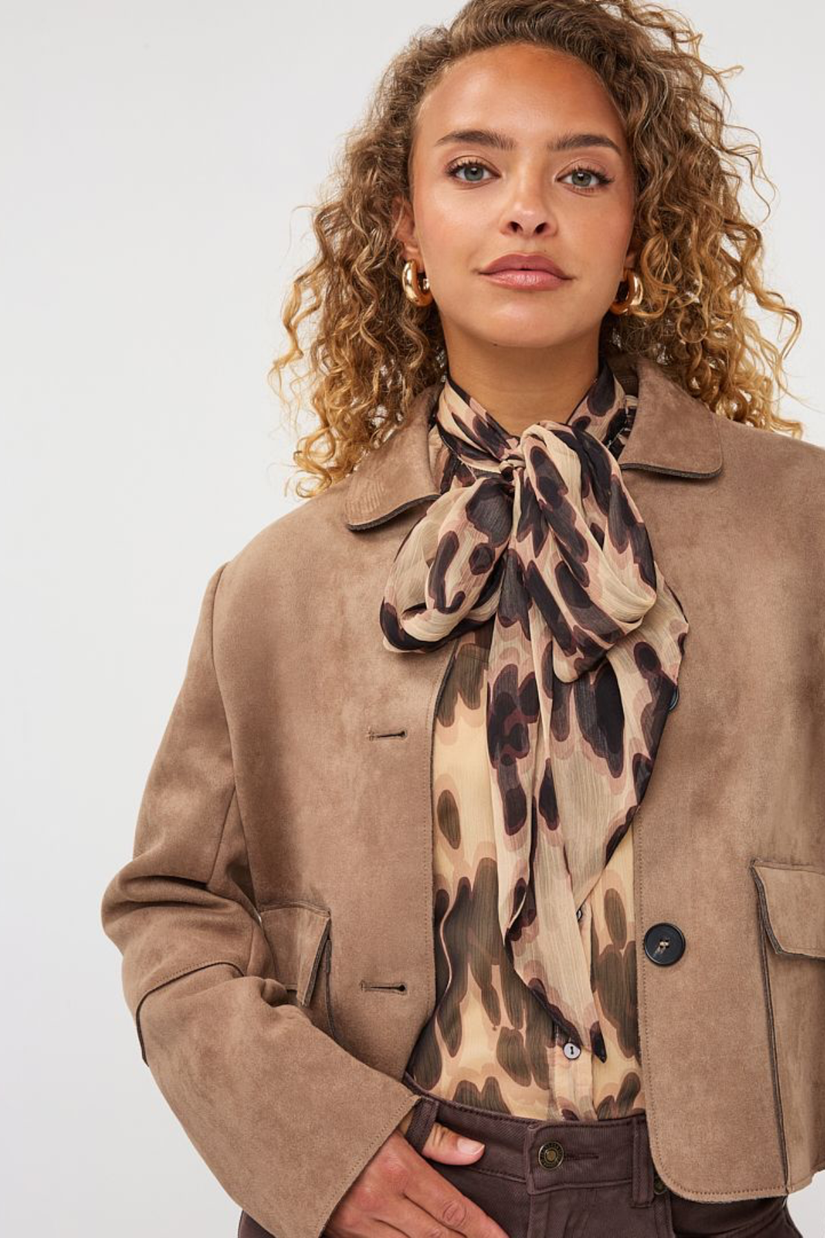 A woman wearing a tan suede jacket, a beige blouse, and a matching leopard-print silk scarf tied around her neck, photographed against a light gray studio background.