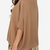 A woman wearing a loose, short-sleeve knit poncho top in a warm camel brown color, paired with tropical print shorts, shown from a side-back angle against a plain light background.