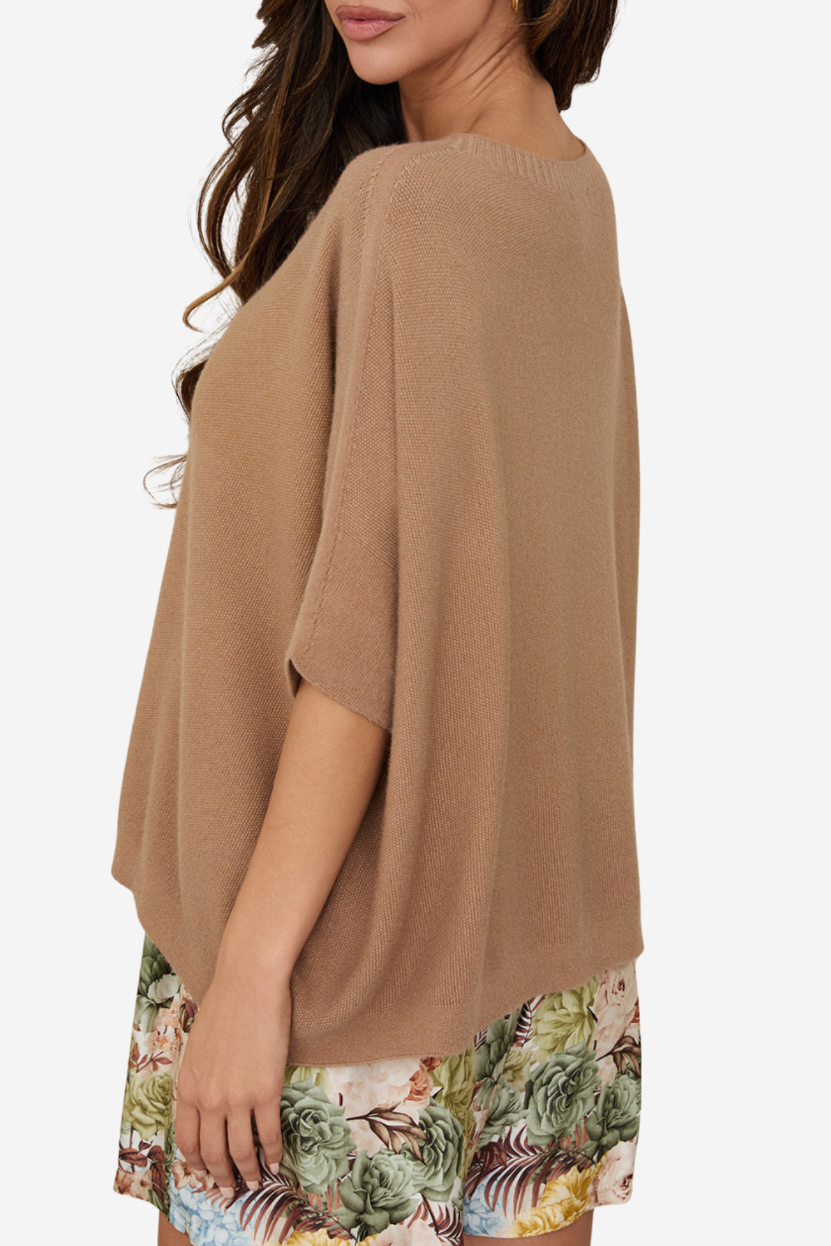 A woman wearing a loose, short-sleeve knit poncho top in a warm camel brown color, paired with tropical print shorts, shown from a side-back angle against a plain light background.