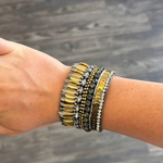 A bohemian-style beaded bracelet on a person's wrist, featuring gold, silver, and dark gray beads, shown against a dark wood floor background.