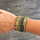 A bohemian-style beaded bracelet on a person's wrist, featuring gold, silver, and dark gray beads, shown against a dark wood floor background.