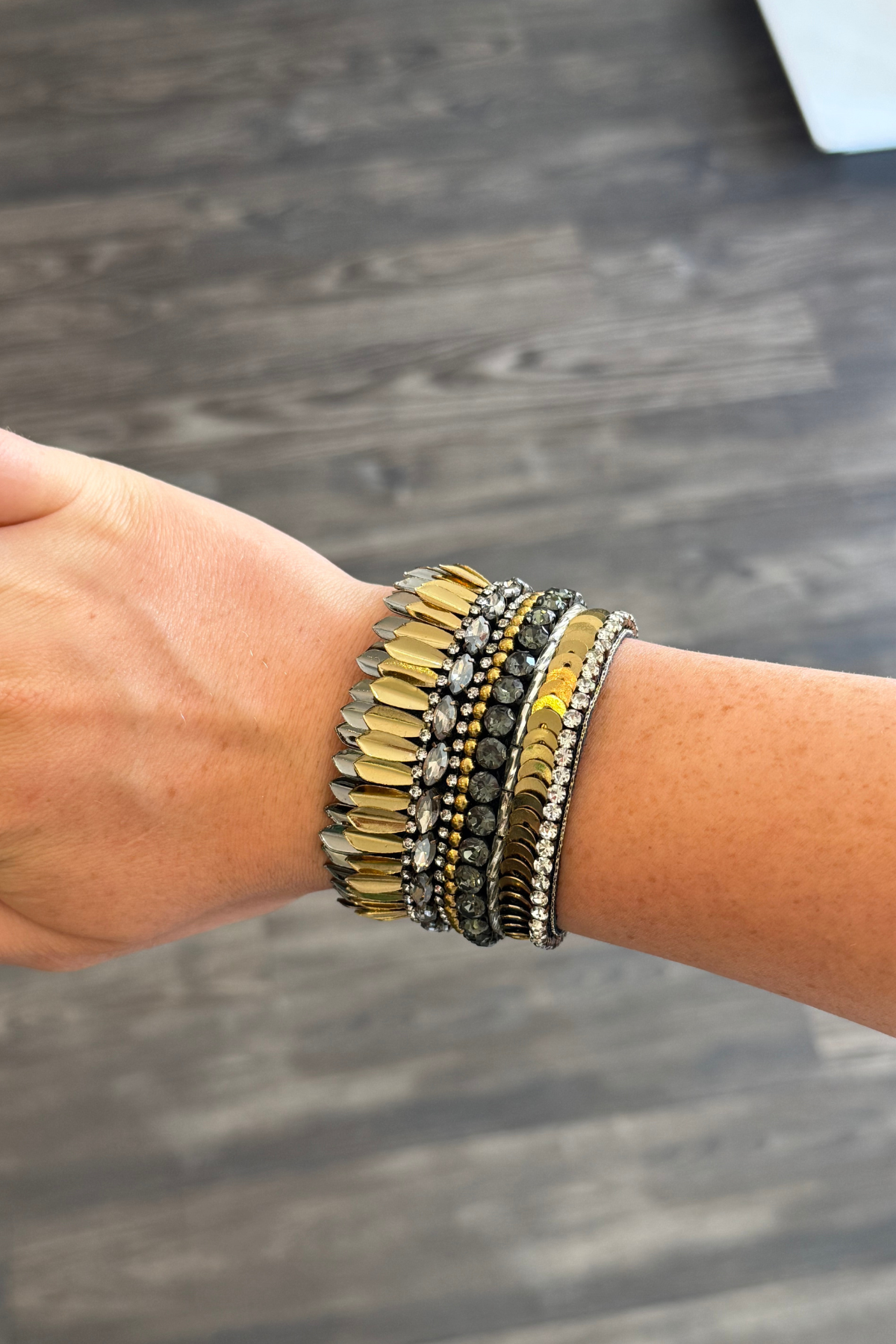 A bohemian-style beaded bracelet on a person's wrist, featuring gold, silver, and dark gray beads, shown against a dark wood floor background.
