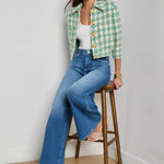 Woman wearing a green patterned jacket and blue jeans sitting on a wooden stool against a white wall.