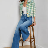 Woman wearing a green patterned jacket and blue jeans sitting on a wooden stool against a white wall.