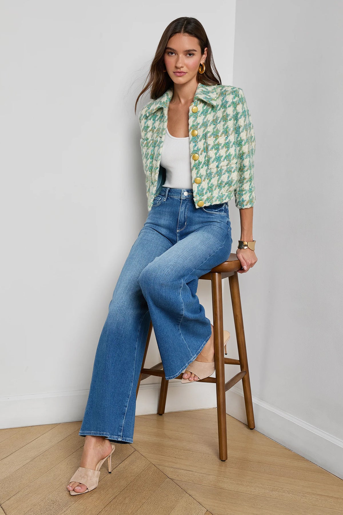 Woman wearing a green patterned jacket and blue jeans sitting on a wooden stool against a white wall.