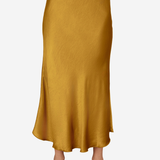 A woman wearing a flowing midi skirt in a warm mustard yellow or ochre color, paired with clear high-heeled sandals, shown against a plain light gray studio background.