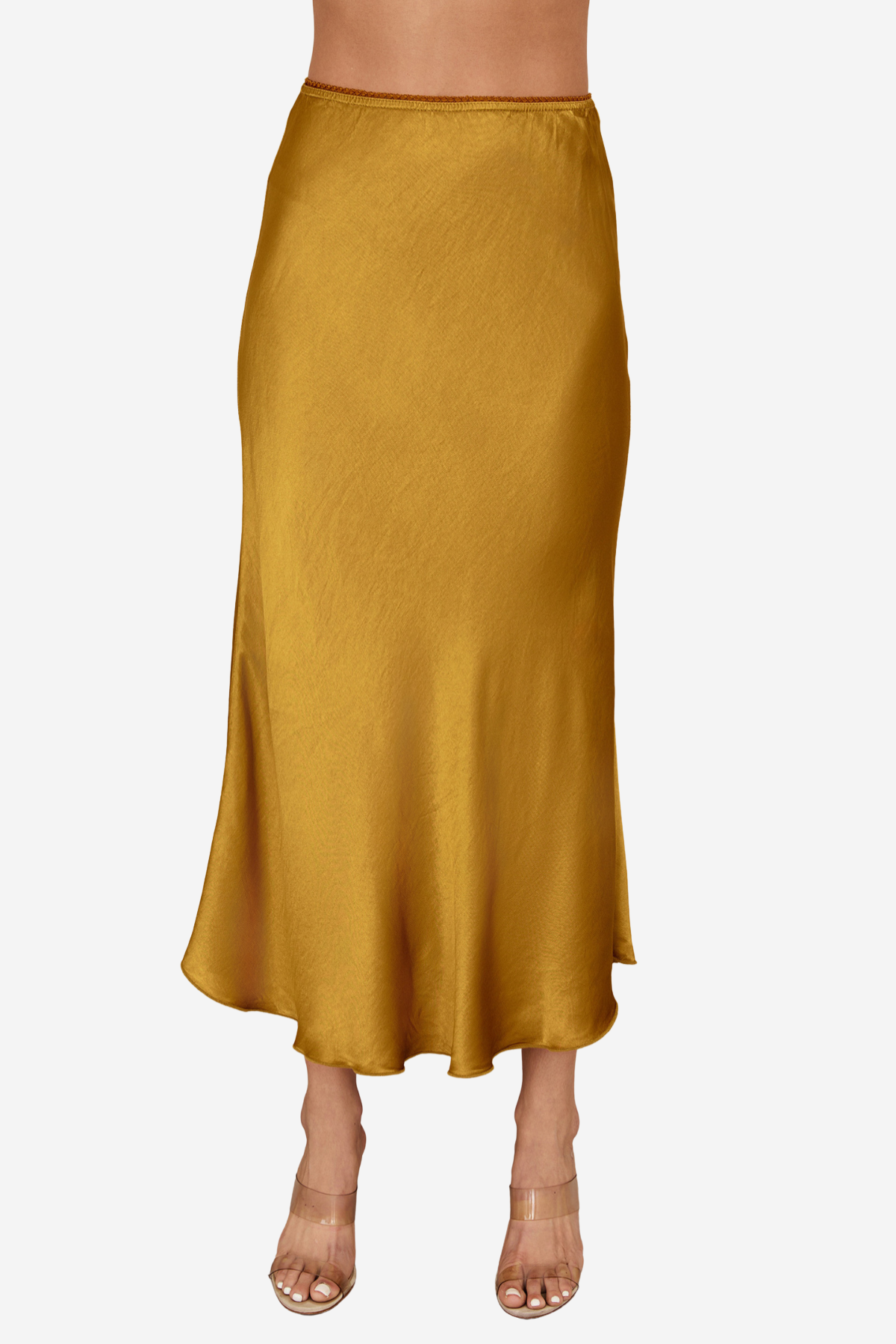 A woman wearing a flowing midi skirt in a warm mustard yellow or ochre color, paired with clear high-heeled sandals, shown against a plain light gray studio background.