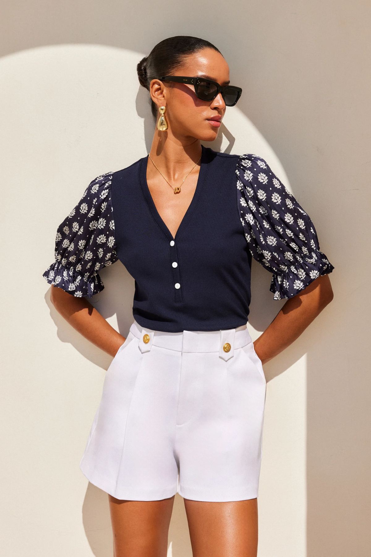 Woman wearing a navy floral blouse and white shorts against a beige wall.