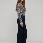 A woman stands in profile wearing a long-sleeve button-up shirt with a dark blue and beige floral pattern, paired with dark wide-leg trousers and brown leather shoes, against a light gray studio background.