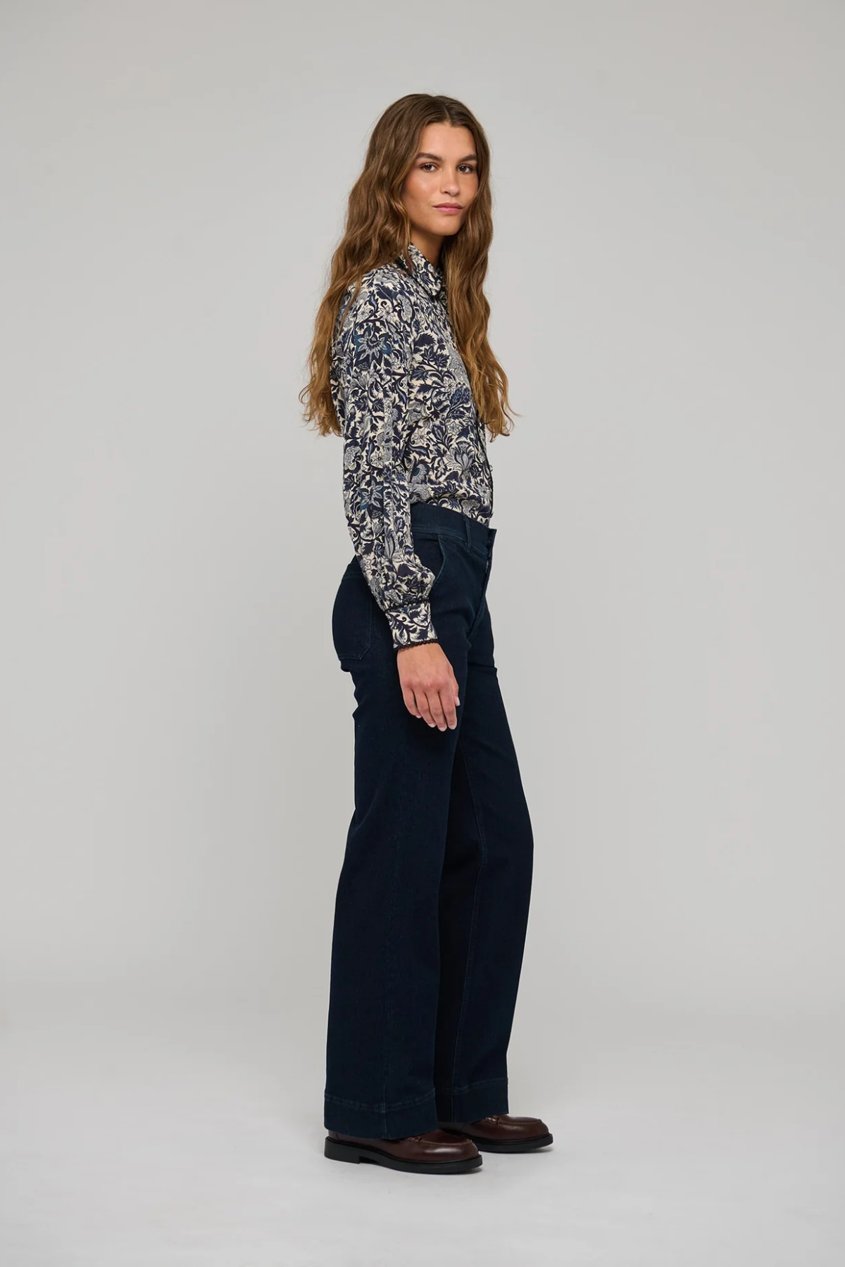 A woman stands in profile wearing a long-sleeve button-up shirt with a dark blue and beige floral pattern, paired with dark wide-leg trousers and brown leather shoes, against a light gray studio background.