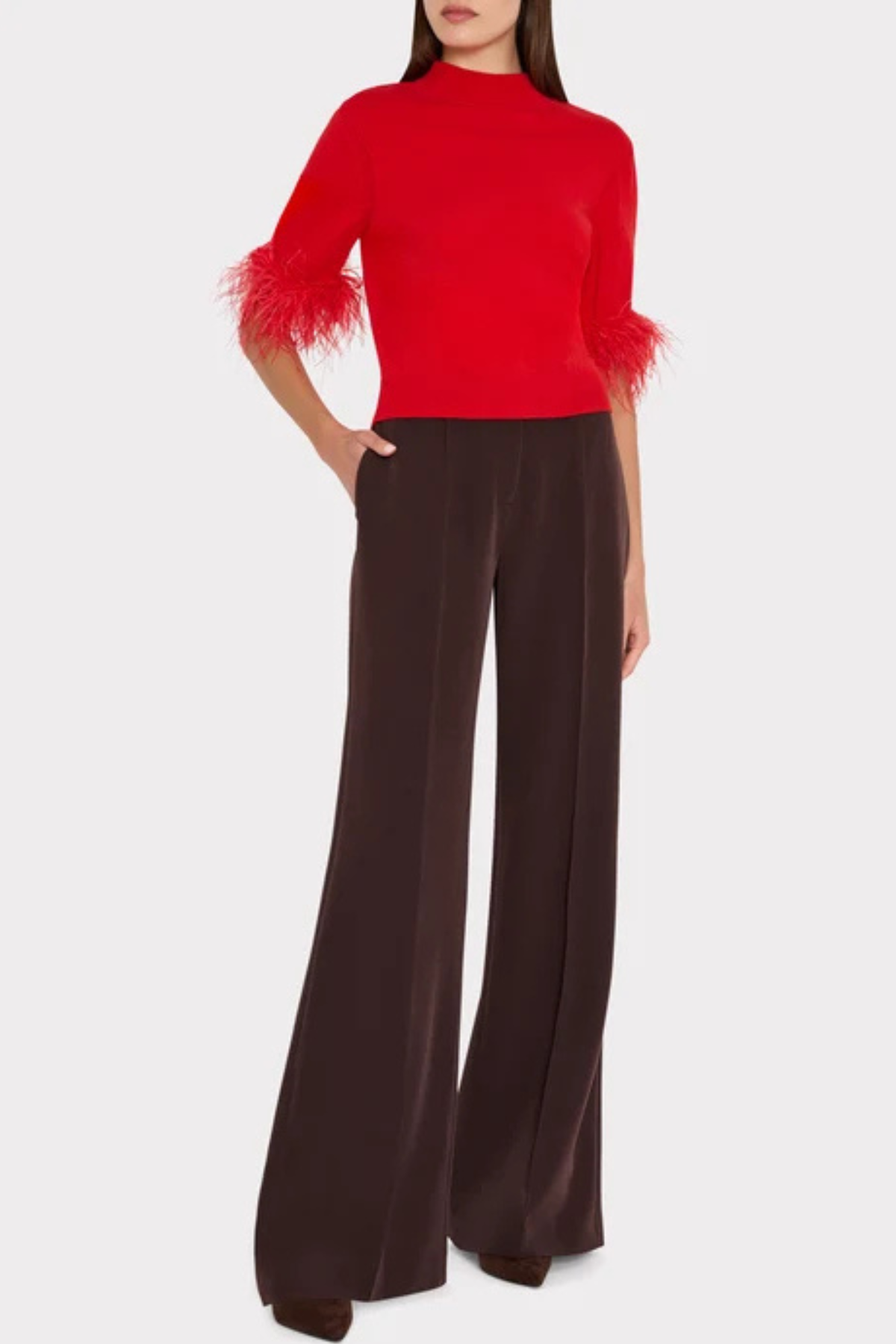A model wearing a vibrant red mock-neck top with feathered elbow-length sleeves and dark brown wide-leg trousers, shown against a light gray studio background.