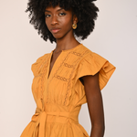 A woman with a large black afro hairstyle wearing a mustard yellow ruffled blouse with a tie waist belt, photographed against a plain light beige background.