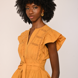 A woman with a large black afro hairstyle wearing a mustard yellow ruffled blouse with a tie waist belt, photographed against a plain light beige background.