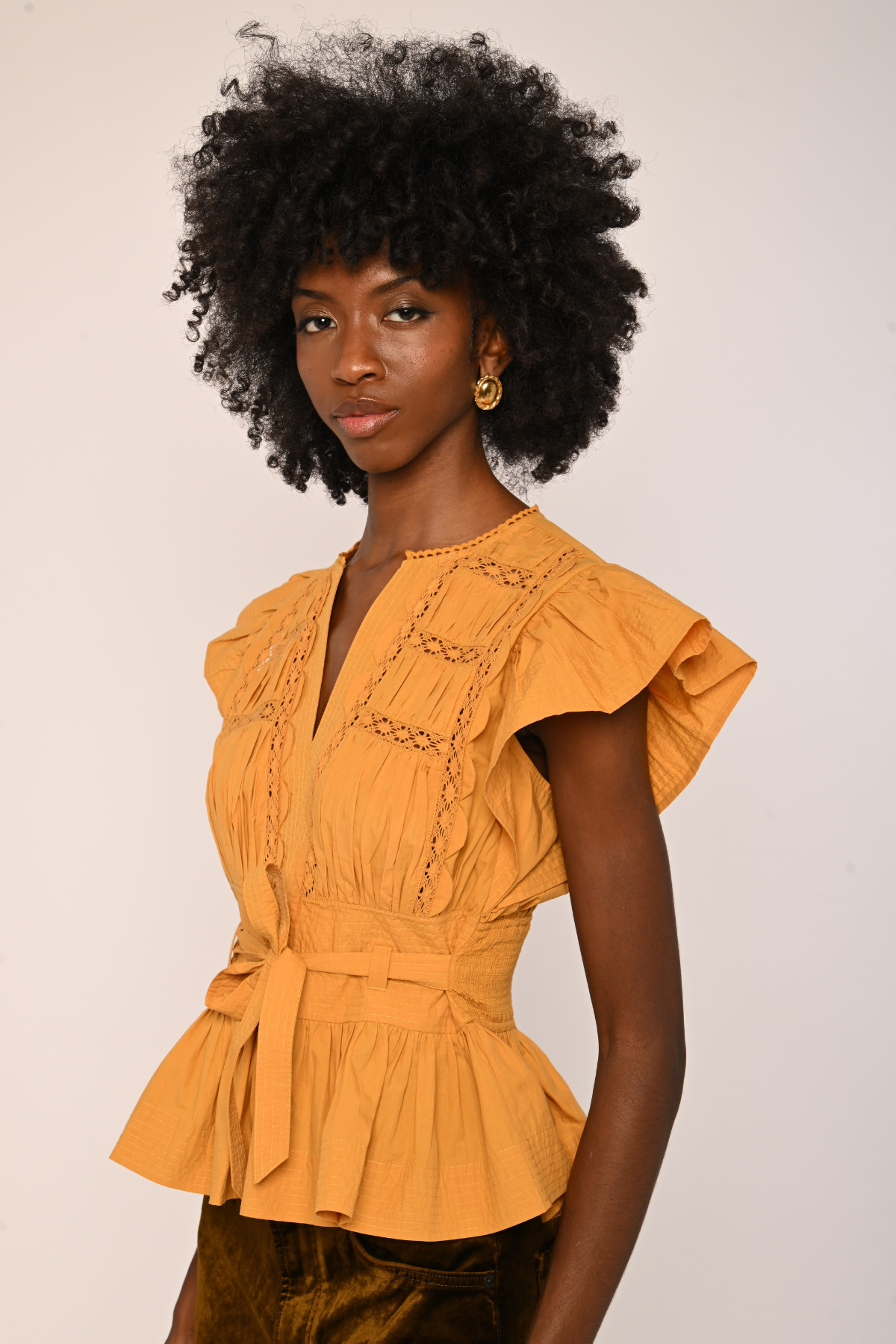 A woman with a large black afro hairstyle wearing a mustard yellow ruffled blouse with a tie waist belt, photographed against a plain light beige background.