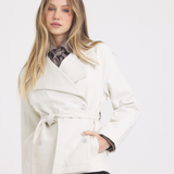 A woman modeling a cream-colored belted wrap jacket over a patterned shirt and blue jeans, set against a plain light gray studio background.
