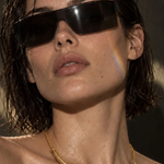 Close-up of a woman wearing oversized black sunglasses and a thin gold collar necklace, with wet hair and warm directional lighting.