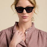 A woman with blonde hair wearing thick black sunglasses and a red-and-white gingham shirt, posed against a solid beige background.