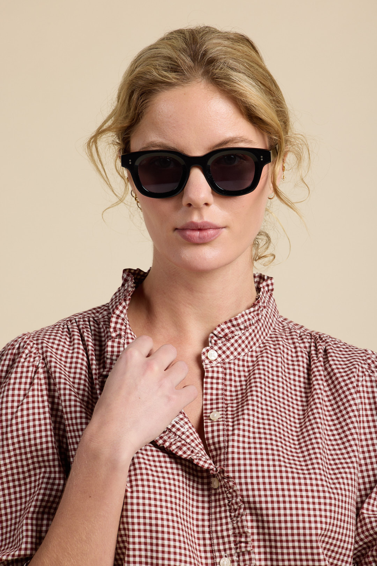 A woman with blonde hair wearing thick black sunglasses and a red-and-white gingham shirt, posed against a solid beige background.