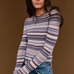 A woman wearing a long-sleeve crewneck knit top with horizontal stripes in purple, blue, and beige, paired with blue jeans and black sunglasses, set against a solid brown background.