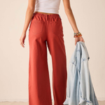 A woman shown from the back wearing rust-colored wide-leg pants, a white tank top, and white sandals, holding a light blue denim jacket against a plain beige wall.