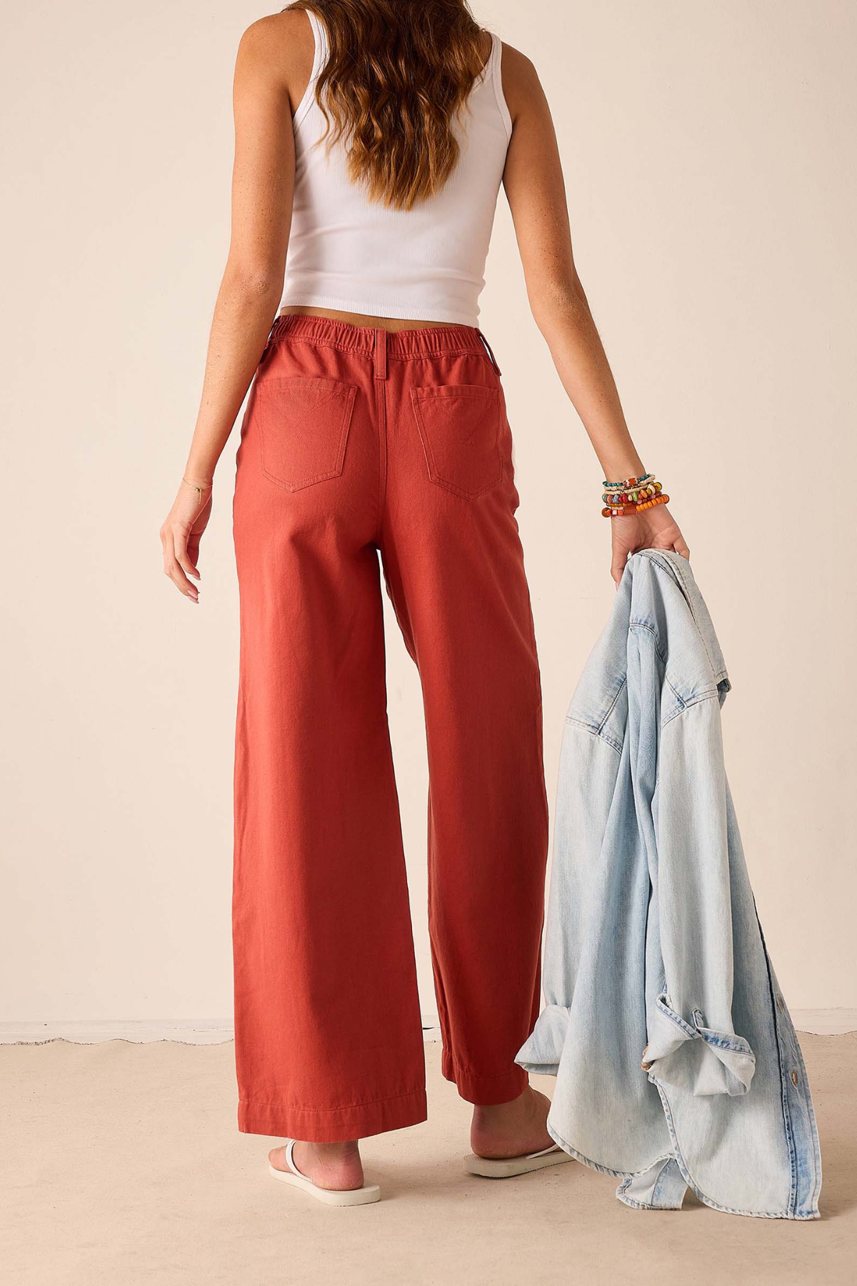 A woman shown from the back wearing rust-colored wide-leg pants, a white tank top, and white sandals, holding a light blue denim jacket against a plain beige wall.