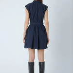 A woman viewed from the back wearing a sleeveless navy dress with thin vertical pinstripes and a matching tie belt, paired with black knee-high heeled boots against a light gray studio background.