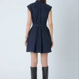 A woman viewed from the back wearing a sleeveless navy dress with thin vertical pinstripes and a matching tie belt, paired with black knee-high heeled boots against a light gray studio background.