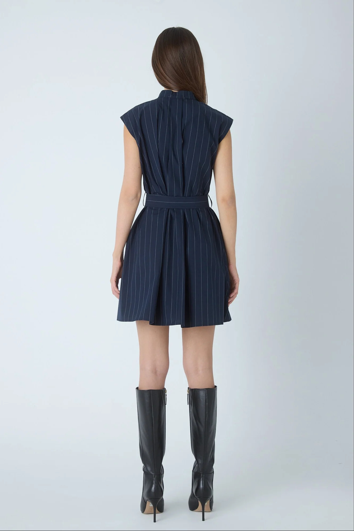 A woman viewed from the back wearing a sleeveless navy dress with thin vertical pinstripes and a matching tie belt, paired with black knee-high heeled boots against a light gray studio background.