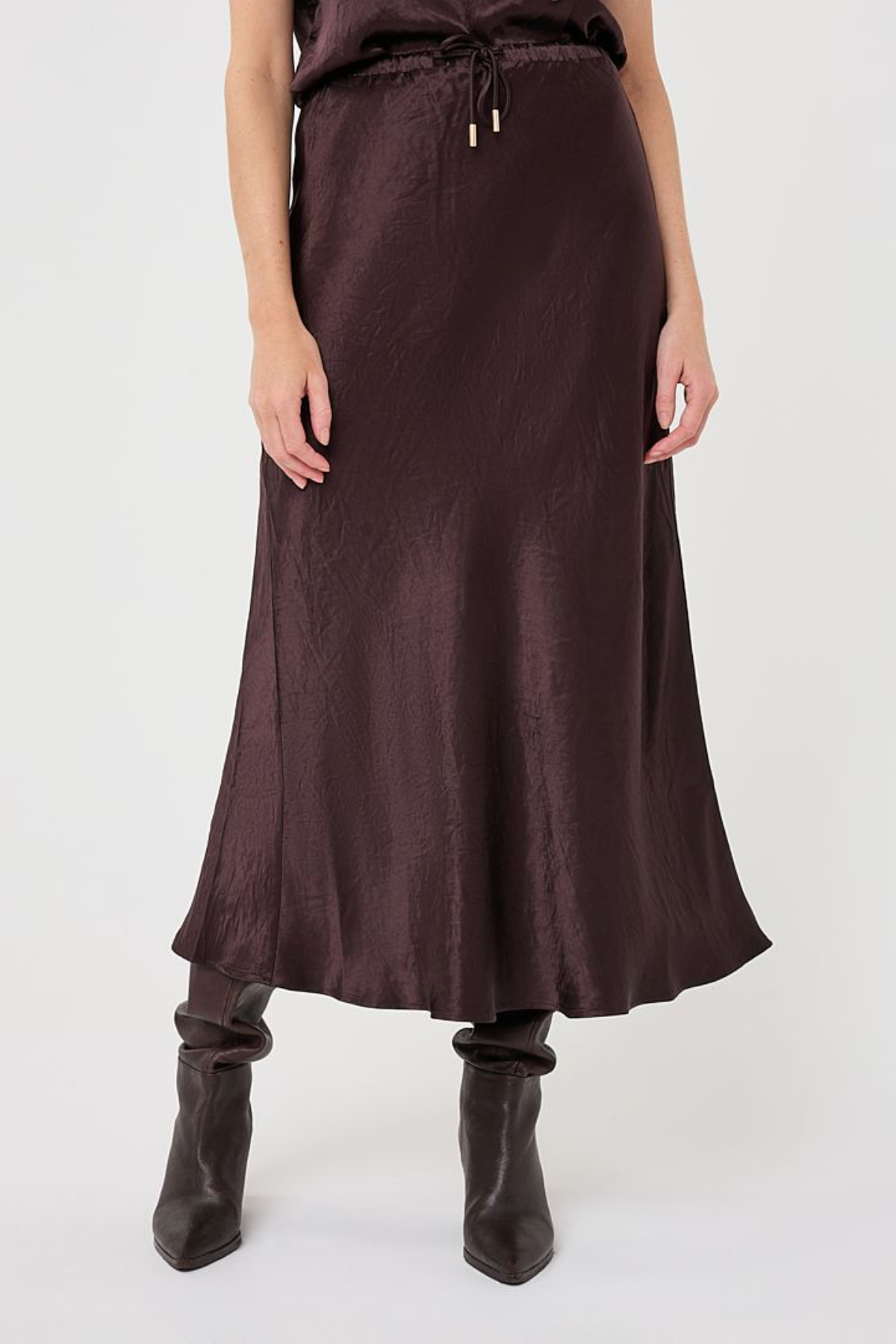 A model wearing a dark brown satin midi skirt with a drawstring waist and dark brown pointed-toe ankle boots, shown from the waist down on a light gray studio background.
