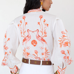 Woman wearing a white blouse with orange floral and bird patterns on a plain background